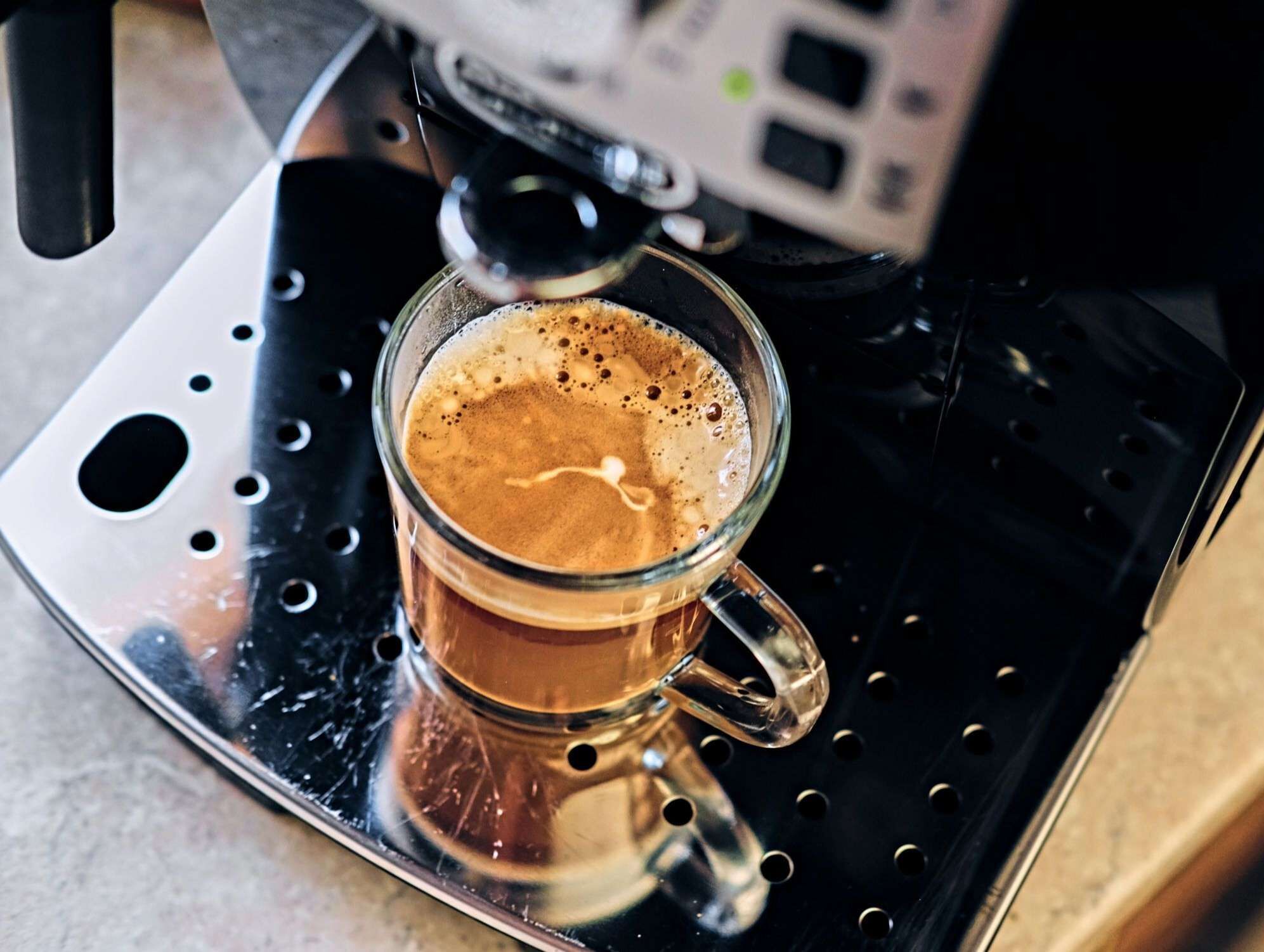Een glazen kop koffie op een metalen dienblad, met een koffieapparaat op de achtergrond.