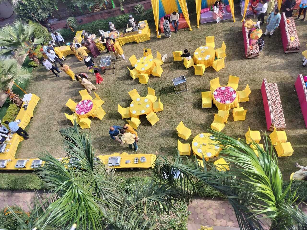 An outdoor event with yellow tables and chairs arranged in a circular pattern, surrounded by greenery and palm trees.