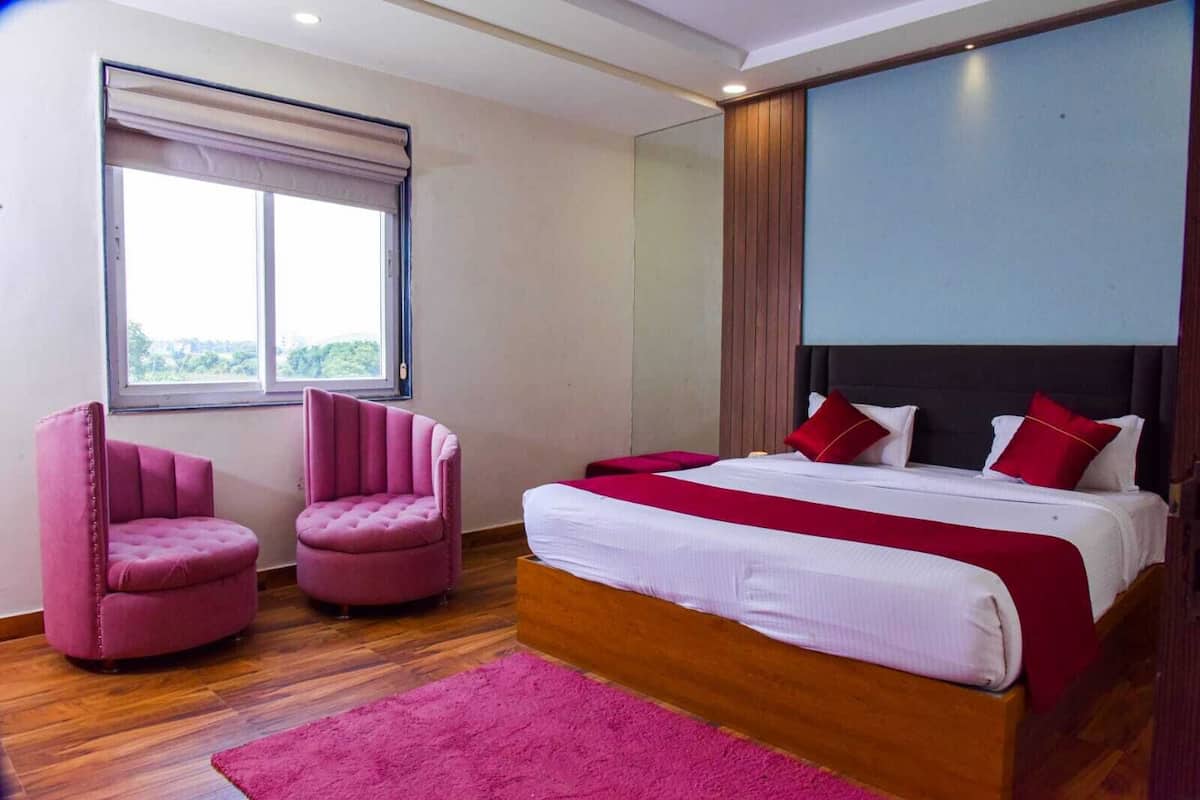 A bedroom with a bed, two pink chairs, a wooden floor, a window, and a view of greenery.