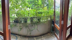A view through a glass door into a room with a white wall and a window overlooking a garden with green foliage. - Cozy luxury room with beautiful views (Battaramulla)