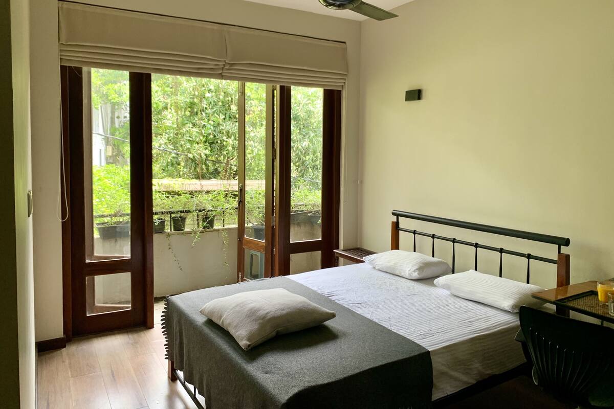 A bedroom with a bed, a ceiling fan, a window with a view of greenery, and a table with a chair.