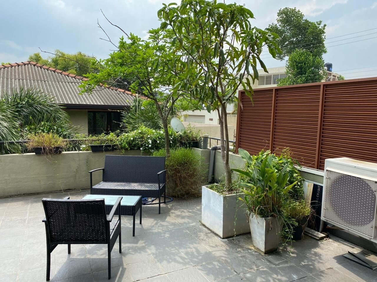 An outdoor patio with a table and chairs, surrounded by potted plants and a water tank in the background.