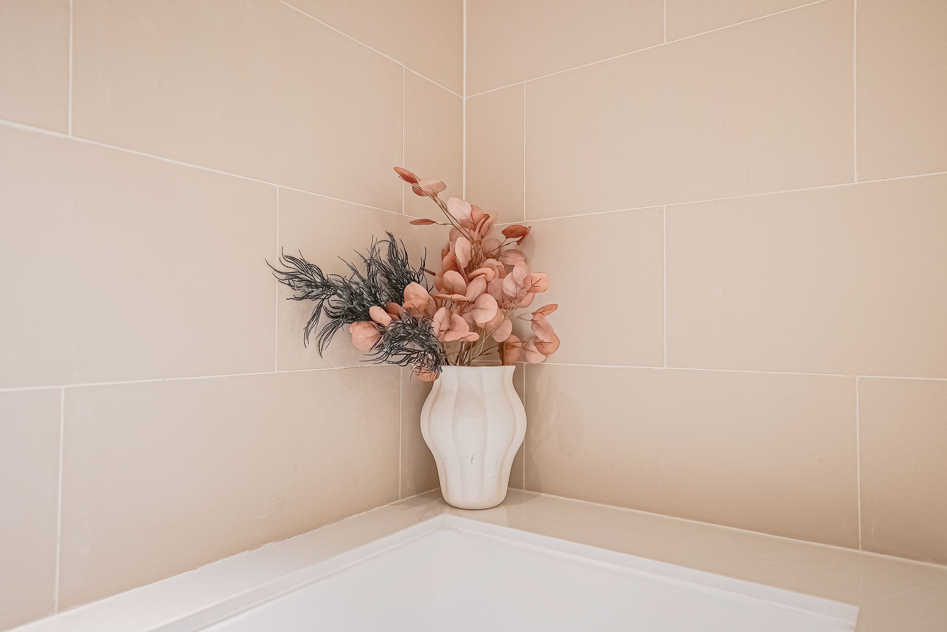 A corner of a bathroom with beige tiles and a white vase with pink flowers and green foliage.