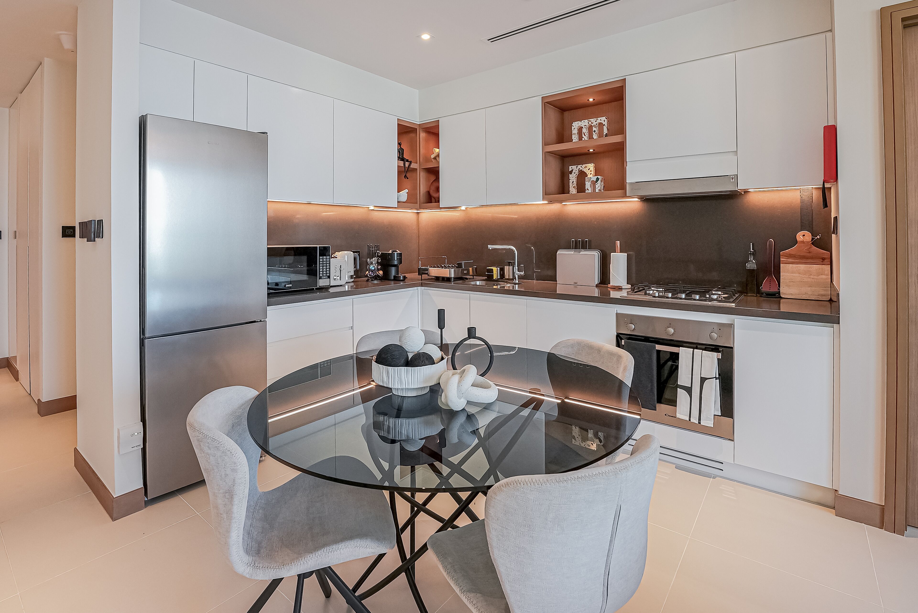 A modern kitchen with white cabinetry, a stainless steel refrigerator, a glass dining table with two chairs, and a built-in oven.