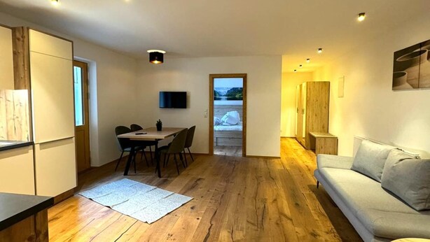 Un salon moderne avec un plancher en bois, un canapé gris, un coin repas comprenant une table et des chaises, et un téléviseur fixé au mur.