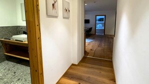Un couloir au plancher de bois mène à une salle à manger équipée d’un téléviseur. Une salle de bain avec un lavabo et des œuvres d’art est visible sur la gauche.