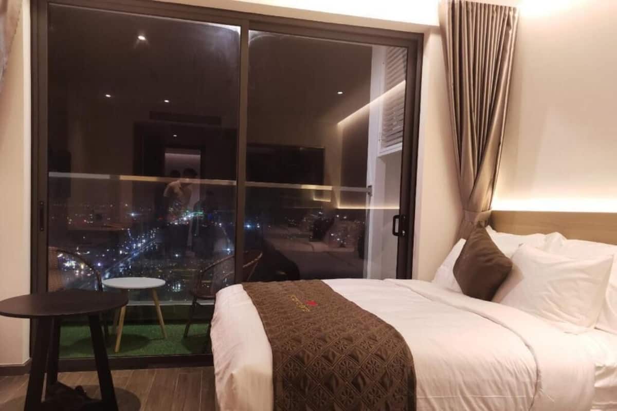 Une chambre d’hôtel avec un grand lit, une petite table et une vue sur le paysage urbain illuminé la nuit.