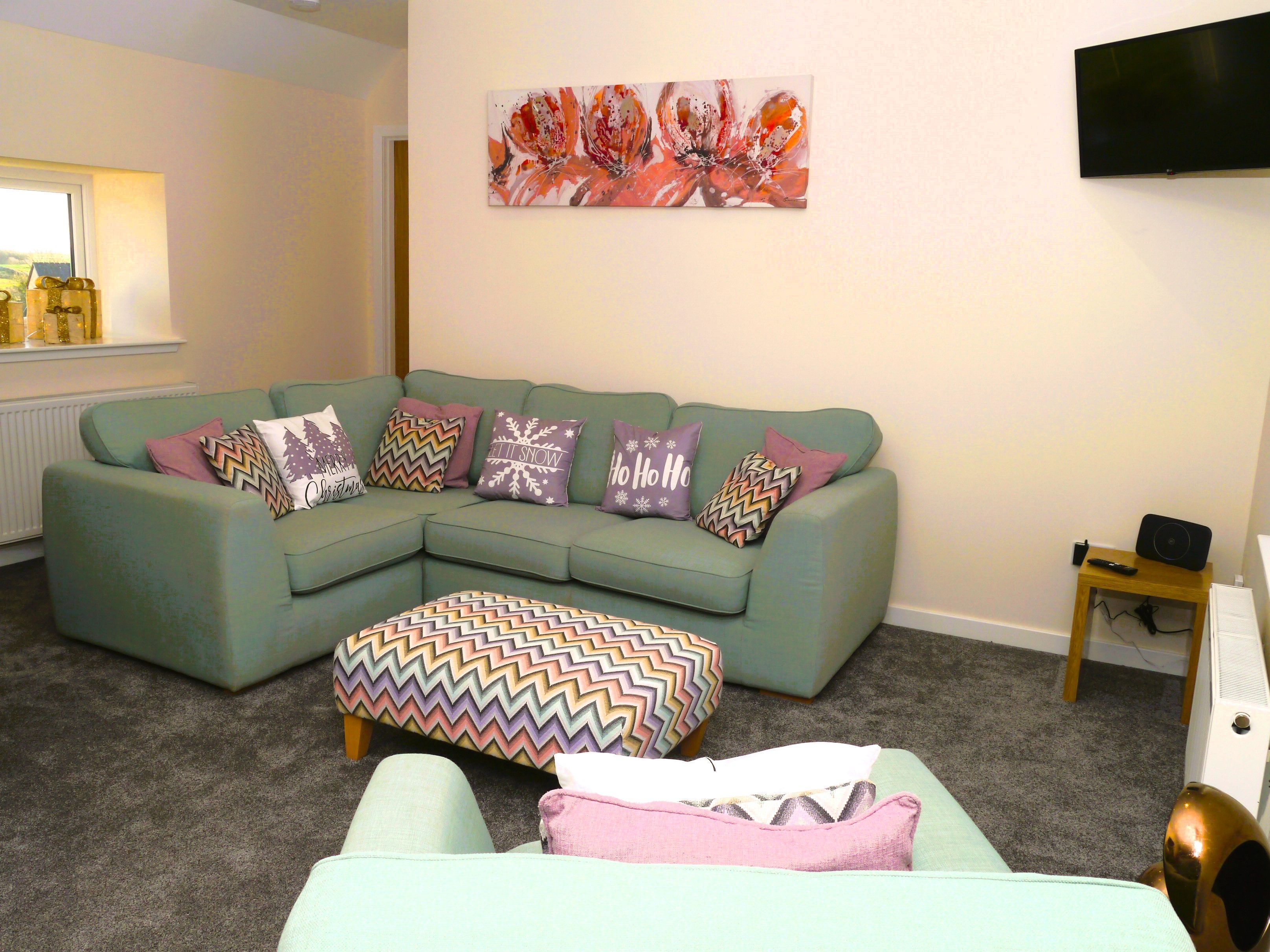 A living room with a sectional sofa, a colorful ottoman, and a flat-screen TV mounted on the wall.