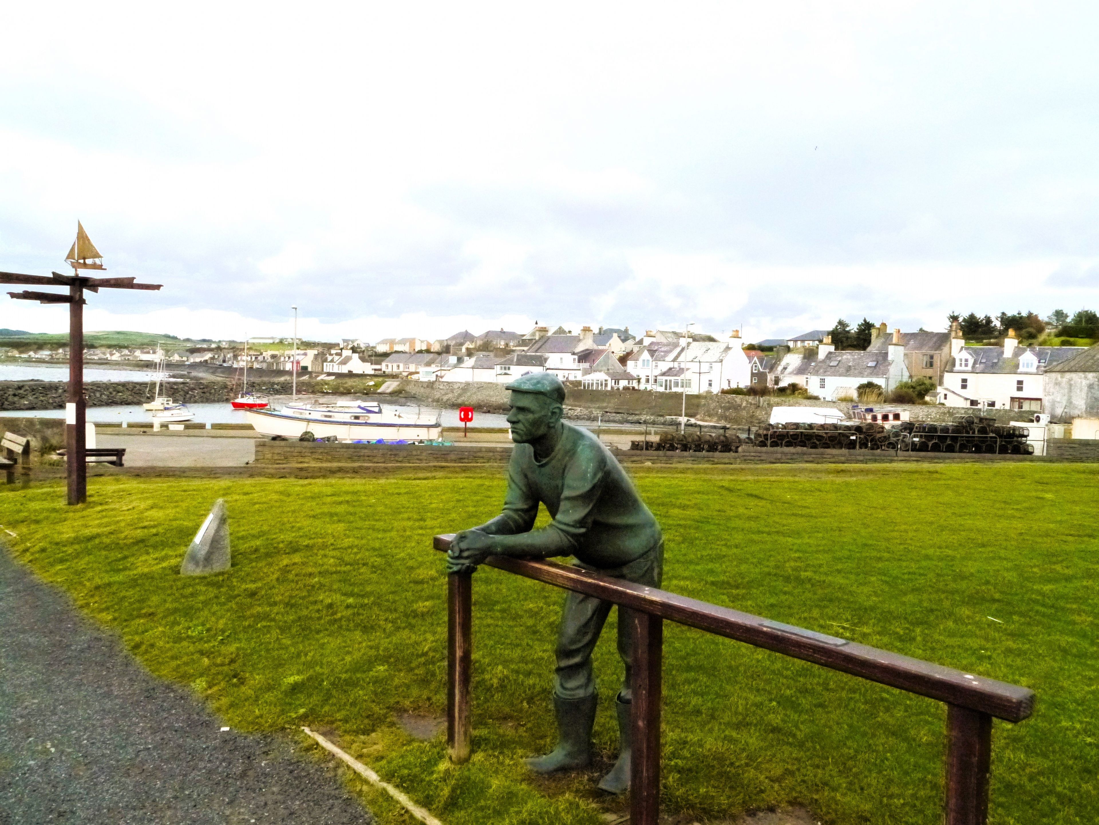 A statue of a man leaning on a railing with a harbor and buildings in the background.