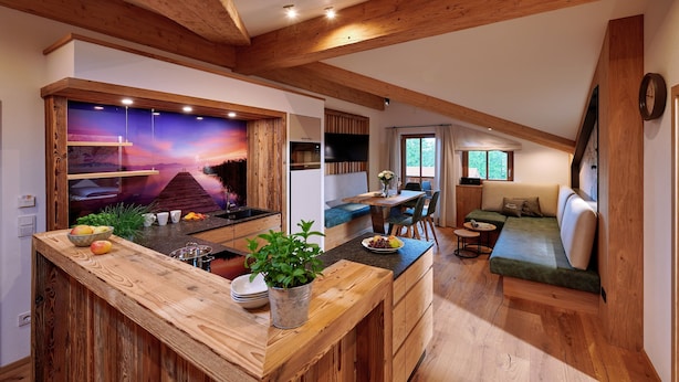 A cozy kitchen with wooden beams, a dining area with a green sofa, and a TV mounted on the wall. - Ferienwohnung Badesteg, 1-5pers., 2sep. SZ, Balkon mit Bergblick (Chieming)
