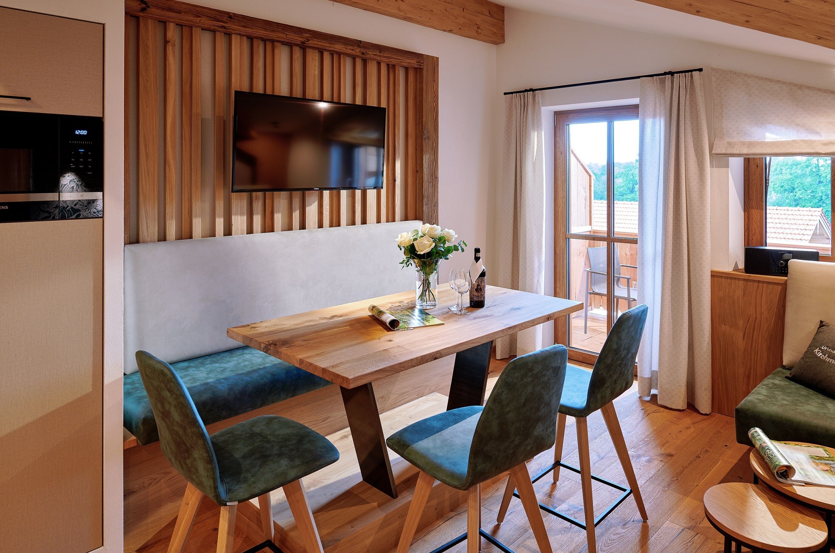 A modern living room with a wooden dining table, green upholstered chairs, a flat-screen TV mounted on a wooden panel, and a view of the outdoors through a large window.