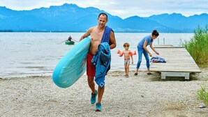 A person carrying a surfboard and a towel, walking on a pebbled beach with a wooden pier and mountains in the background. - Ferienwohnung Bootshütte, 1-5pers., 2sep. SZ, 2.og, Balkon mit Chiemseeblick (Chieming)