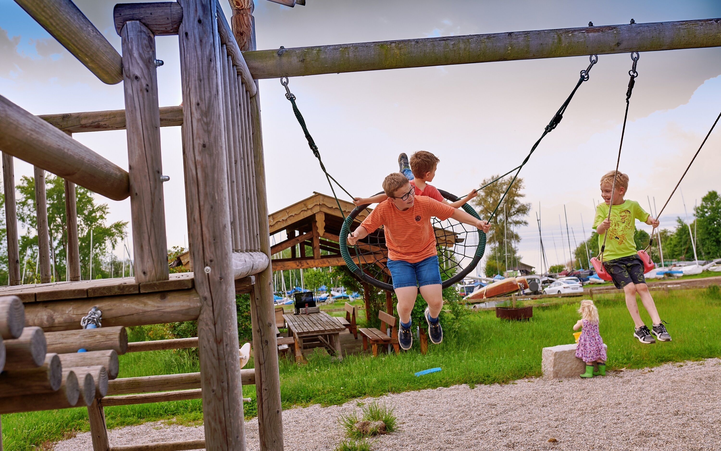 Ein hölzerner Spielplatz mit einer Schaukel mit Netz, einer Holzbank und einem Picknicktisch im Hintergrund.