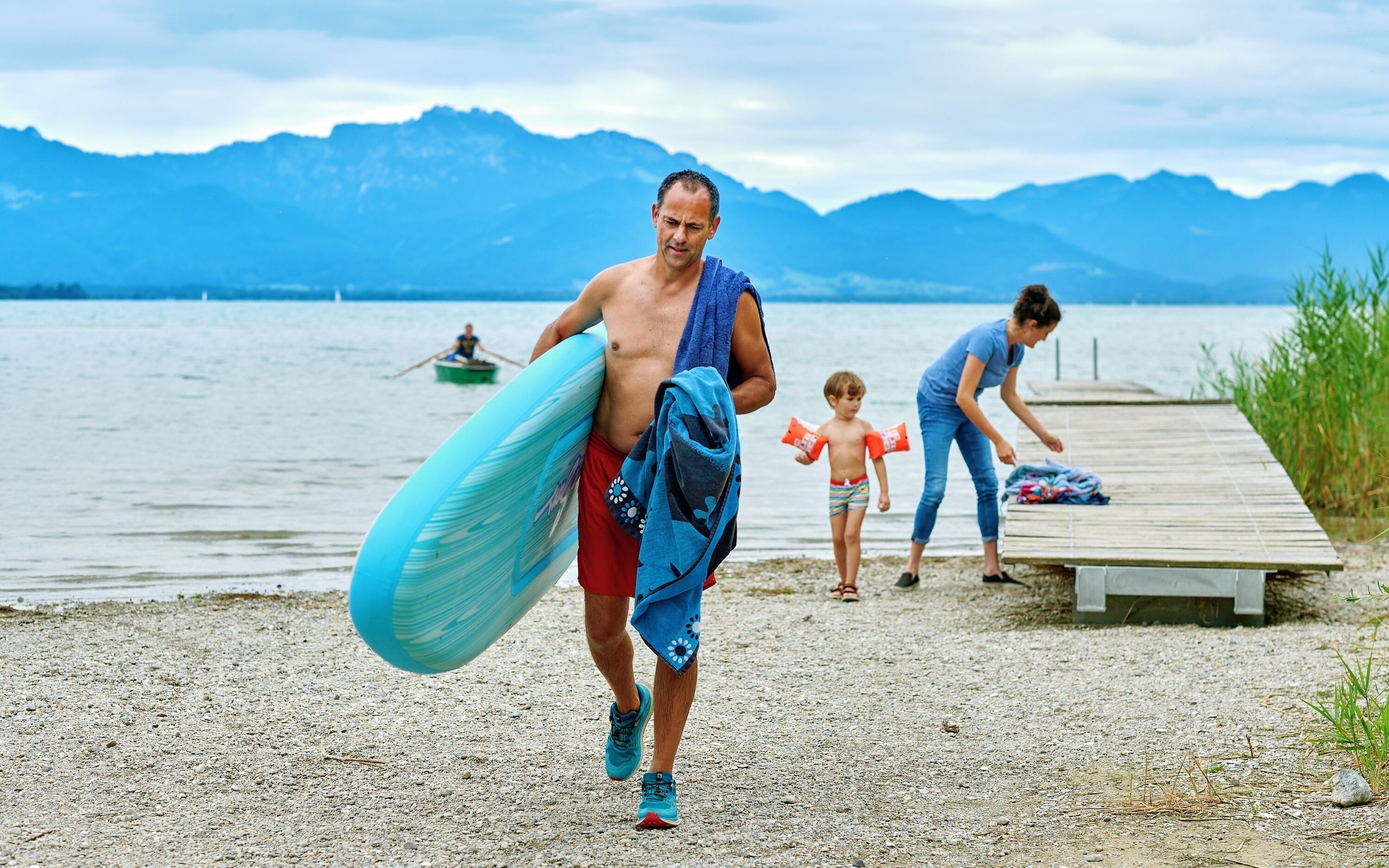 A person carrying a surfboard and a towel, walking on a pebbled beach with a wooden pier and mountains in the background.