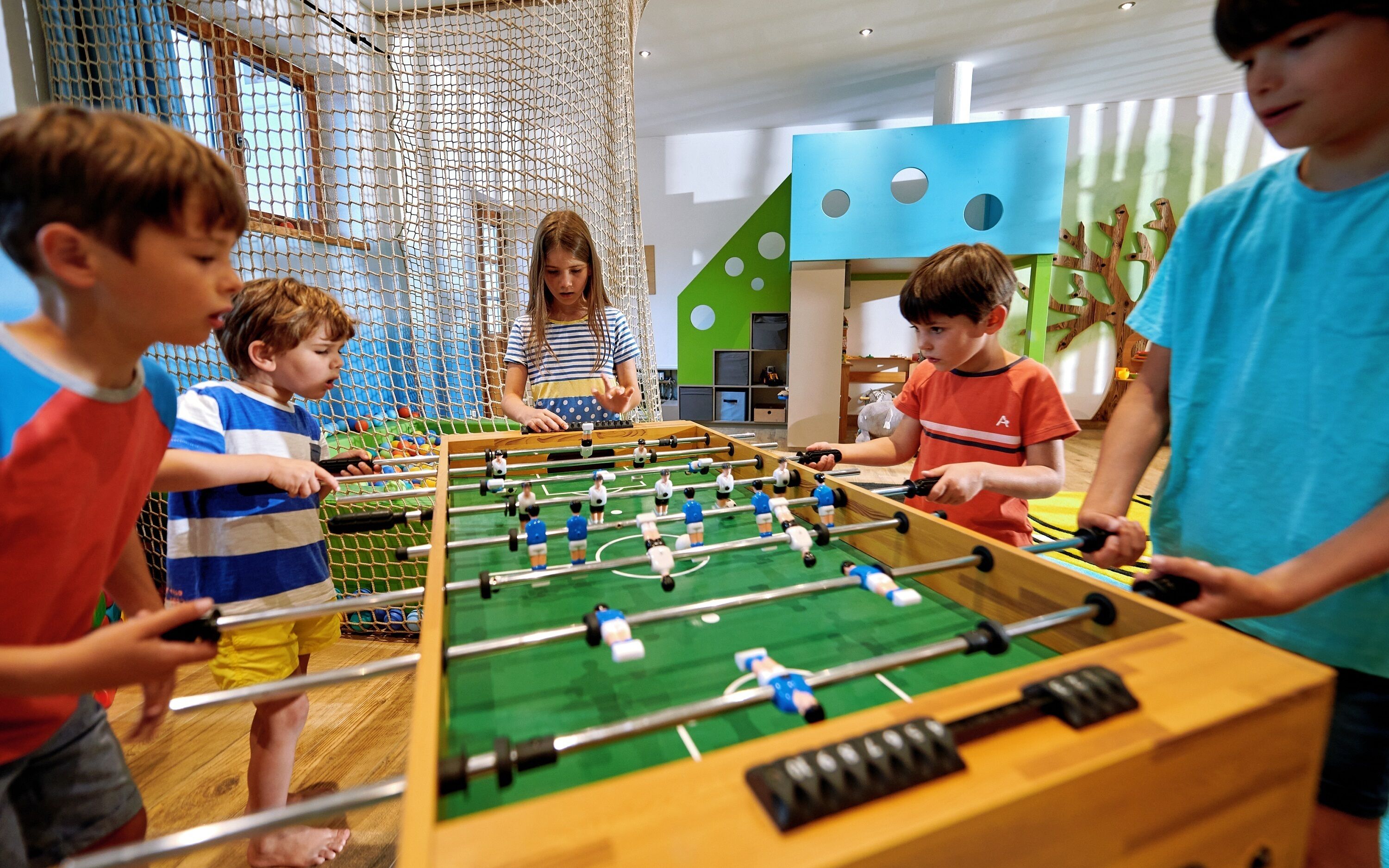 A group of children playing foosball in a brightly lit indoor play area with netted climbing structures and a colorful play wall.