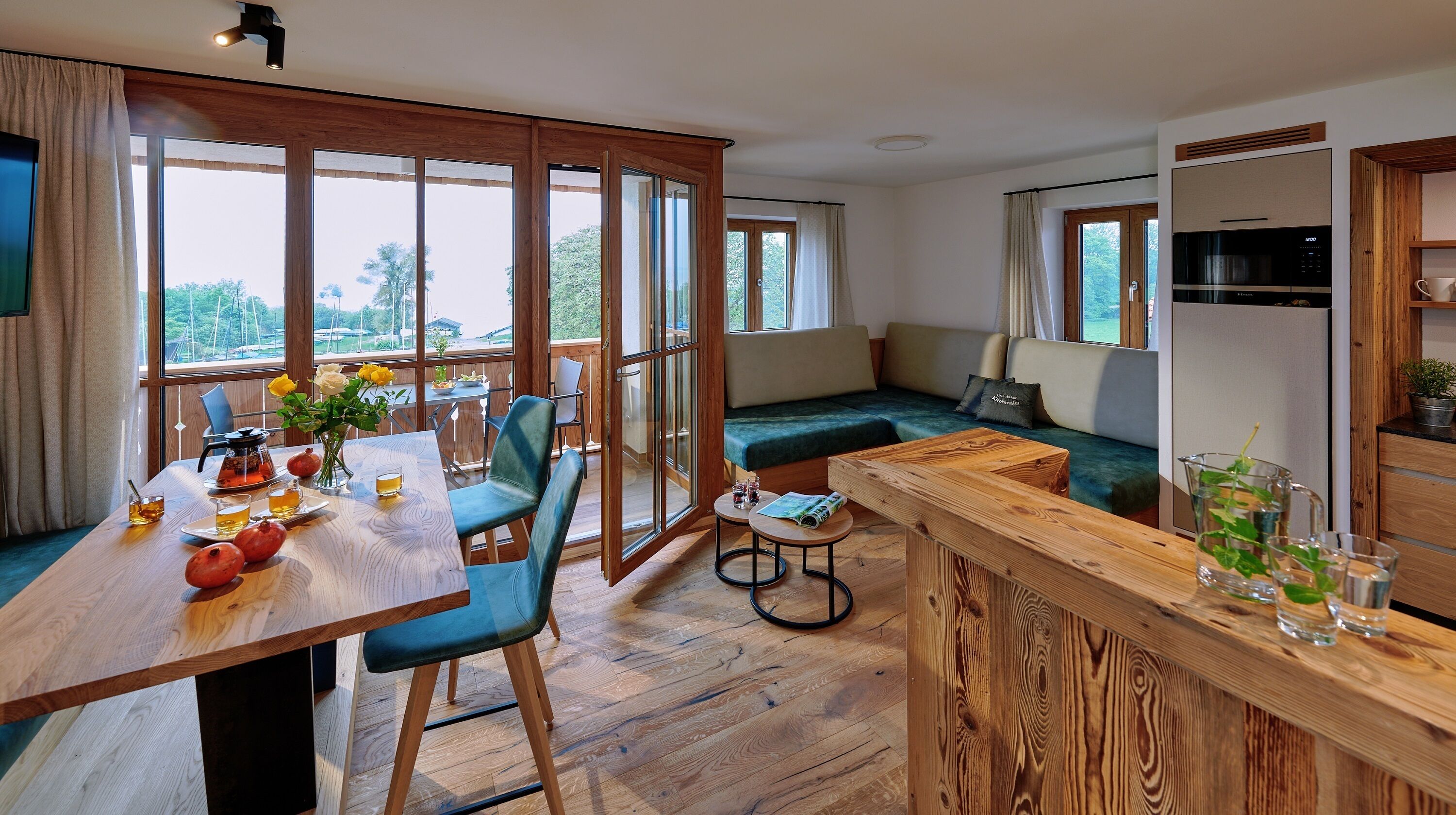 Un salón comedor moderno de planta abierta, con suelo de madera, una mesa de comedor con vasos y una tetera, un sofá con cojines y una cocina con microondas y cafetera.