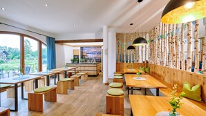 Ein modernes Restaurant mit Holztischen und -bänken, einer Küchenzeile und großen Fenstern mit Blick ins Freie.