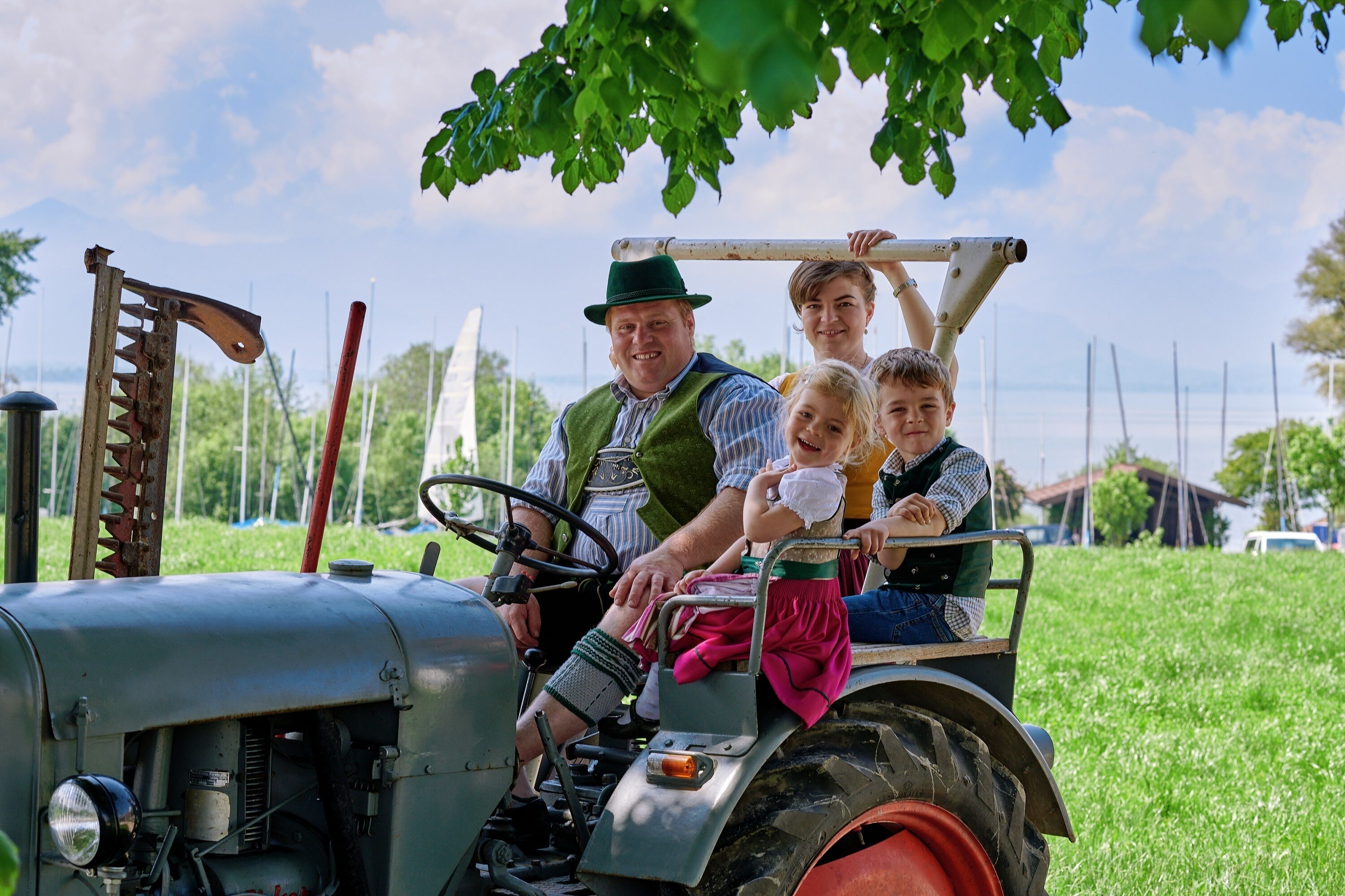 Un vieux tracteur, avec une famille à bord, est garé dans un champ herbeux, entouré d’arbres, avec un ciel dégagé en arrière-plan.