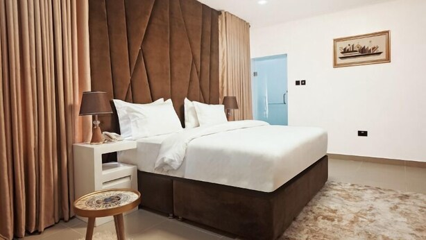 A modern bedroom with a large bed, a nightstand, a round side table, and a headboard with a tufted design. - 3H Villas Hotel & Luxury Apartment (Lekki)