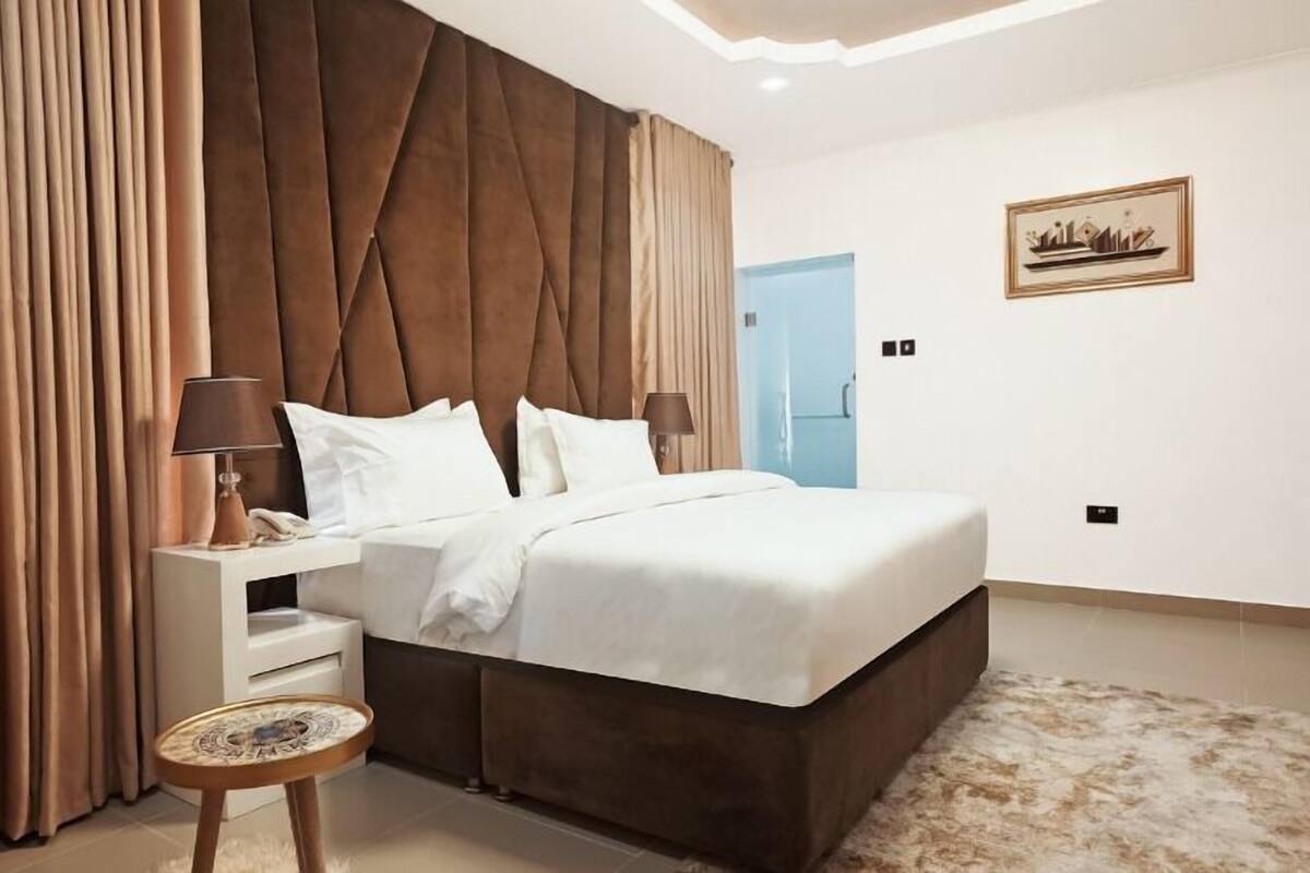 Une chambre à coucher moderne avec un grand lit, une table de chevet, une petite table d’appoint ronde et une tête de lit au motif capitonné.