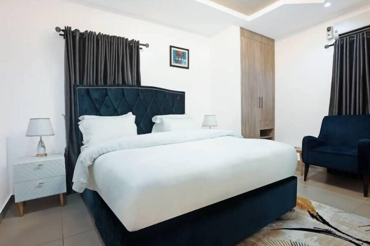 Une chambre d’hôtel moderne avec un grand lit, deux tables de chevet avec des lampes, une armoire et un fauteuil bleu.