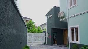 A narrow alleyway with a gated entrance, a building with a dark facade, and a small garden area with pink flowers. - 3H Villas Hotel & Luxury Apartment (Lekki)