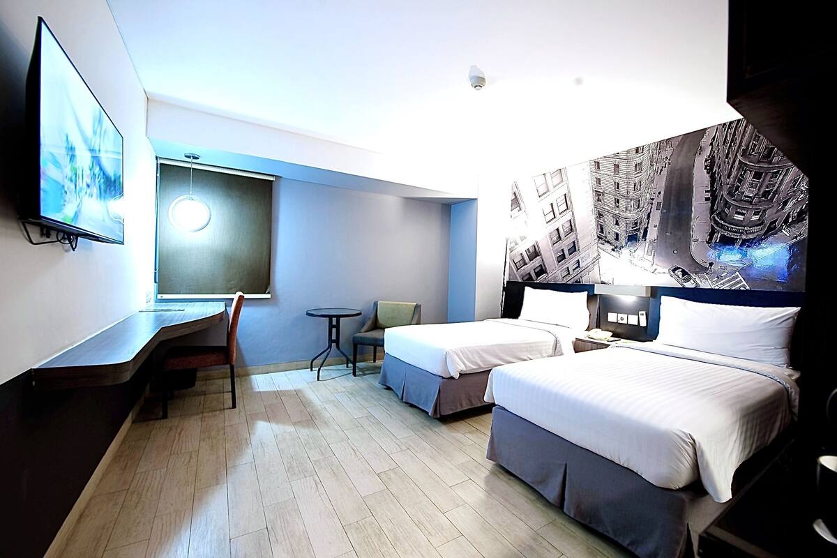A modern hotel room with two beds, a desk, a chair, a small table, and a TV.