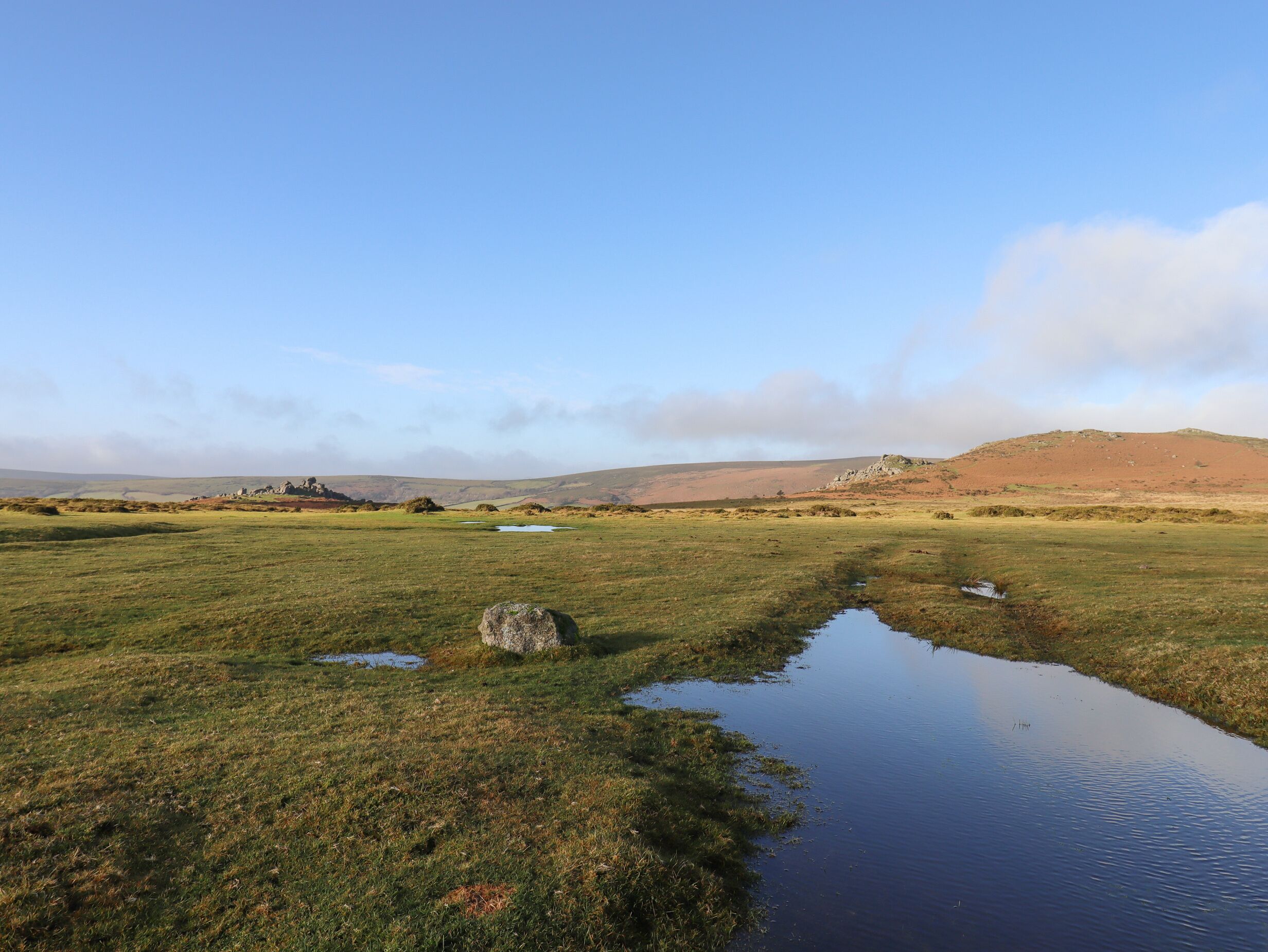 A natural landscape with a small pond, grassy fields, and distant hills.