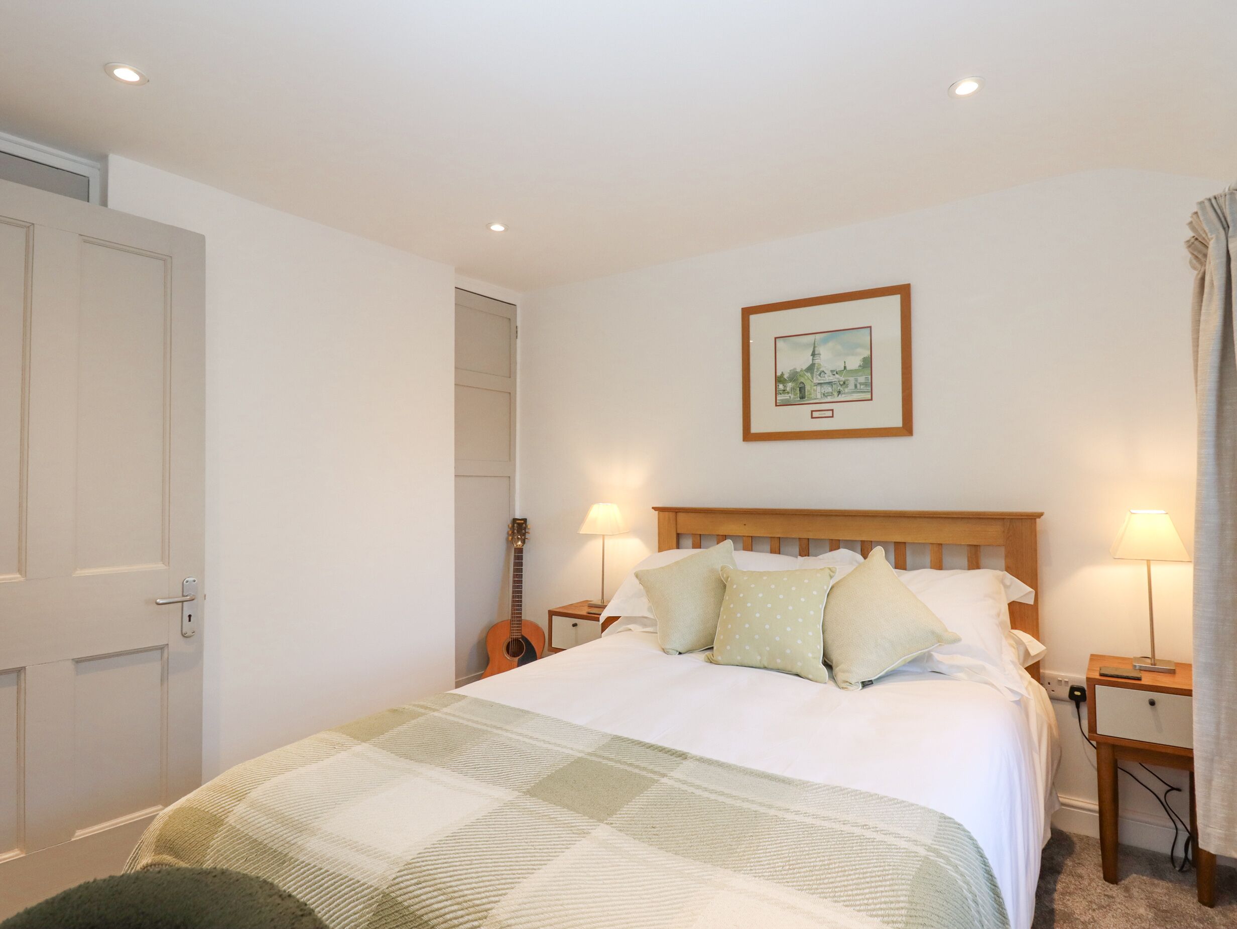 A bedroom with a wooden bed, two bedside lamps, a guitar, a framed picture on the wall, and a door.