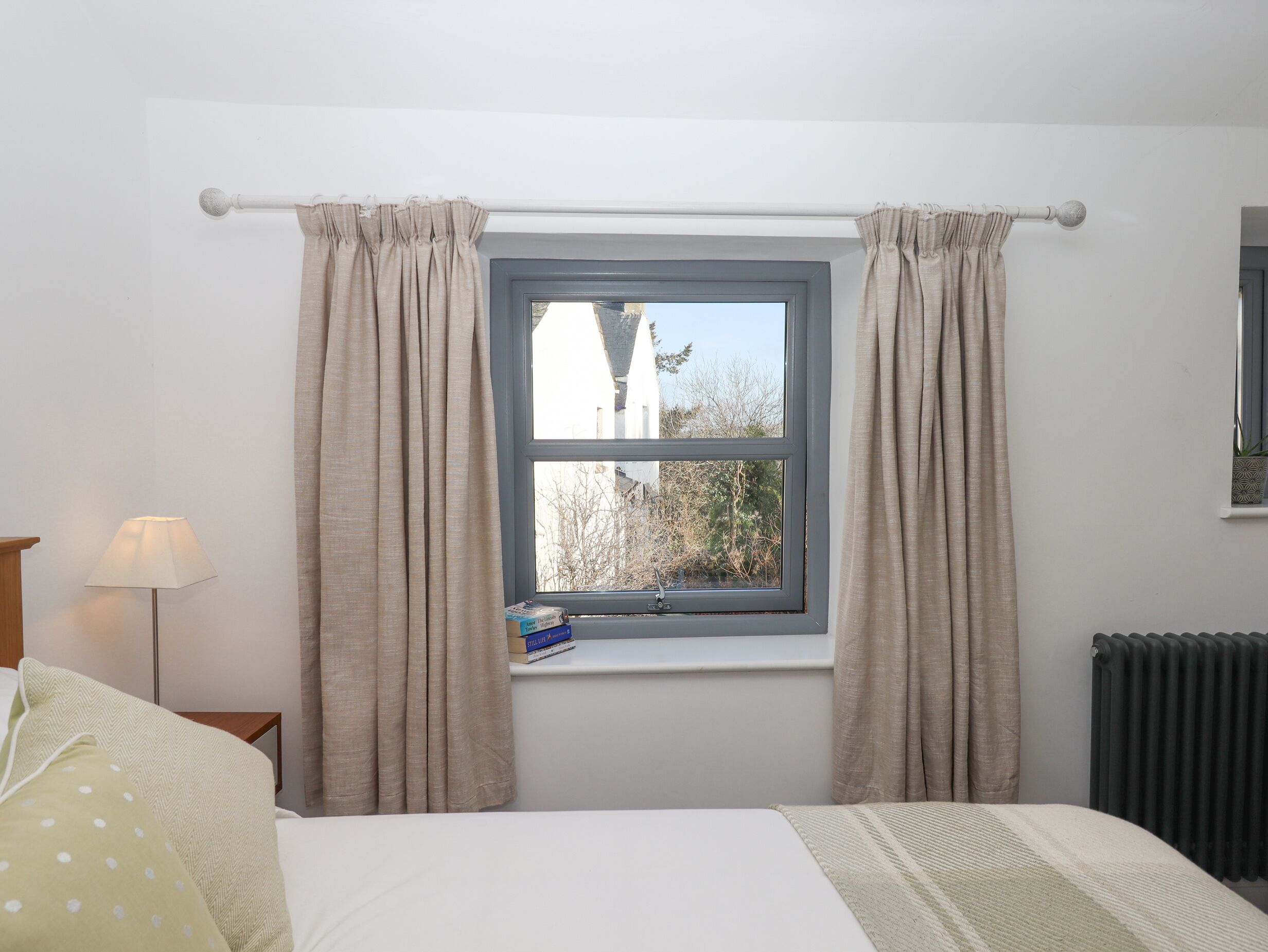 Un dormitorio con ventana, cortinas, lámpara de noche, radiador y cama con manta.