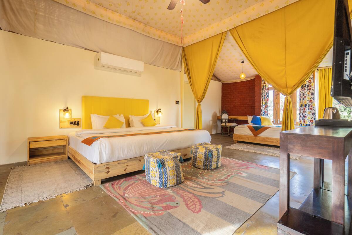Une chambre à coucher comprenant un lit, des tables de chevet, une petite table en bois, une lampe de chevet, une fenêtre avec des rideaux et un téléviseur posé sur un meuble.