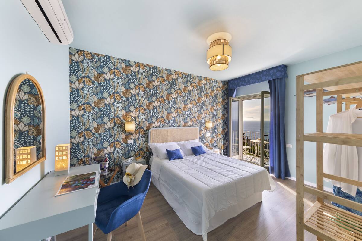 Ein Schlafzimmer mit Bett, Schreibtisch, Stuhl, Spiegel und Blick ins Freie.