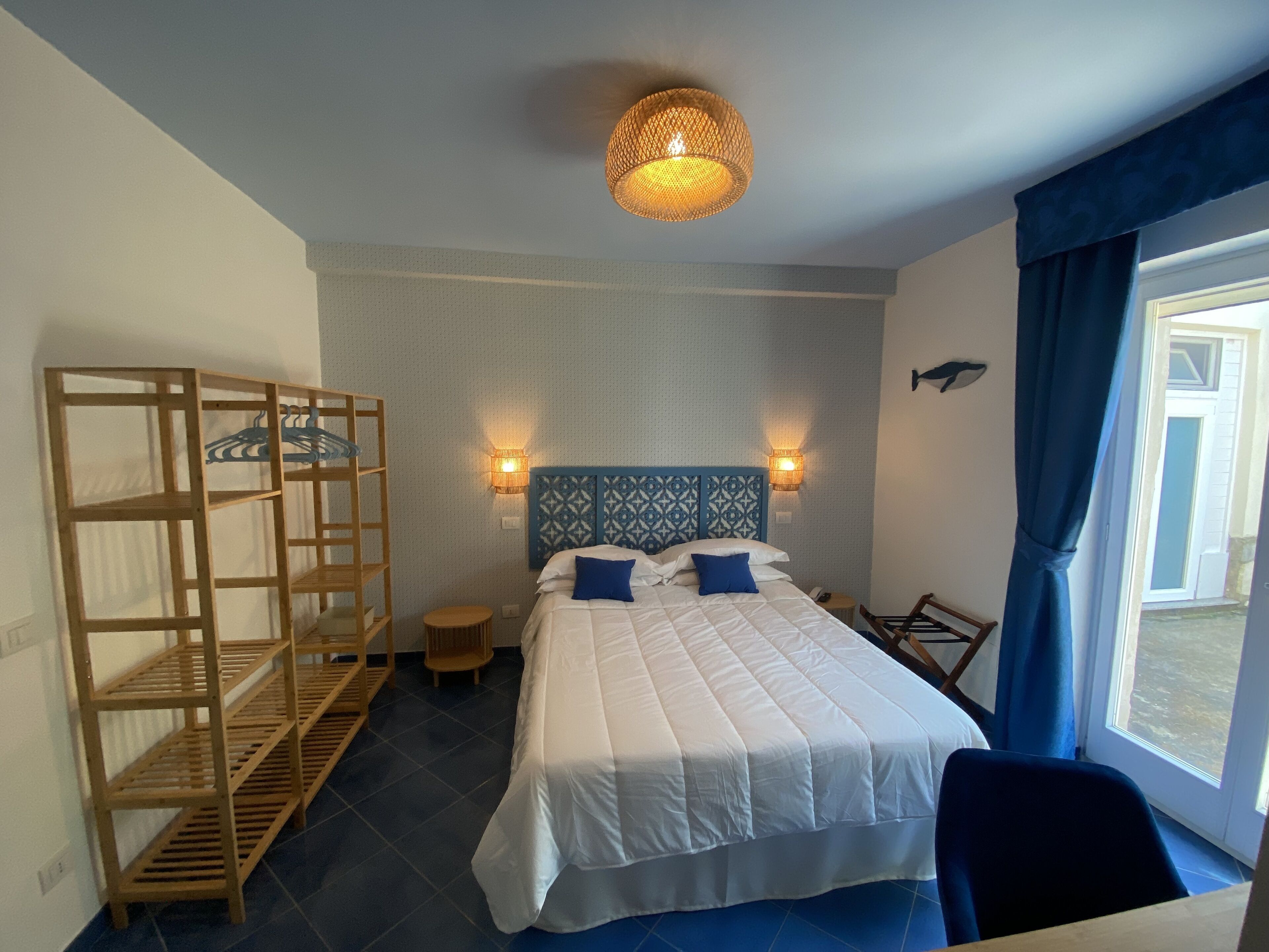 A bedroom with a bed, a wooden shelf unit, two wall-mounted lamps, and a window with curtains.