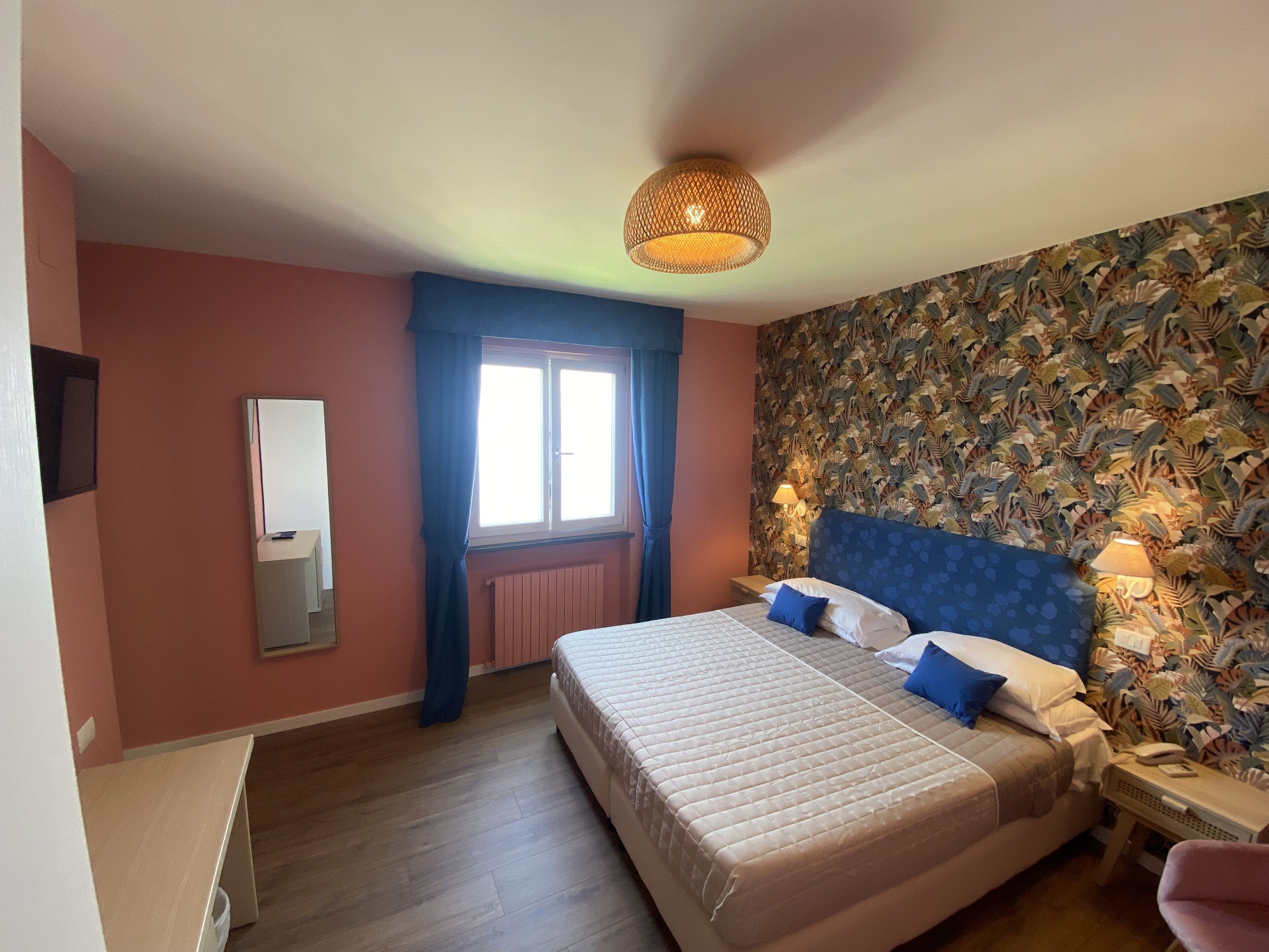 A bedroom with a bed, a nightstand, a lamp, a window with curtains, a radiator, and a wall with a leaf pattern.