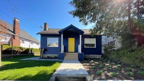 A two-story house with a blue exterior, a yellow front door, and a small front yard with a concrete pathway leading to the entrance. - Skyline Blue House in Greenwood (Seattle)