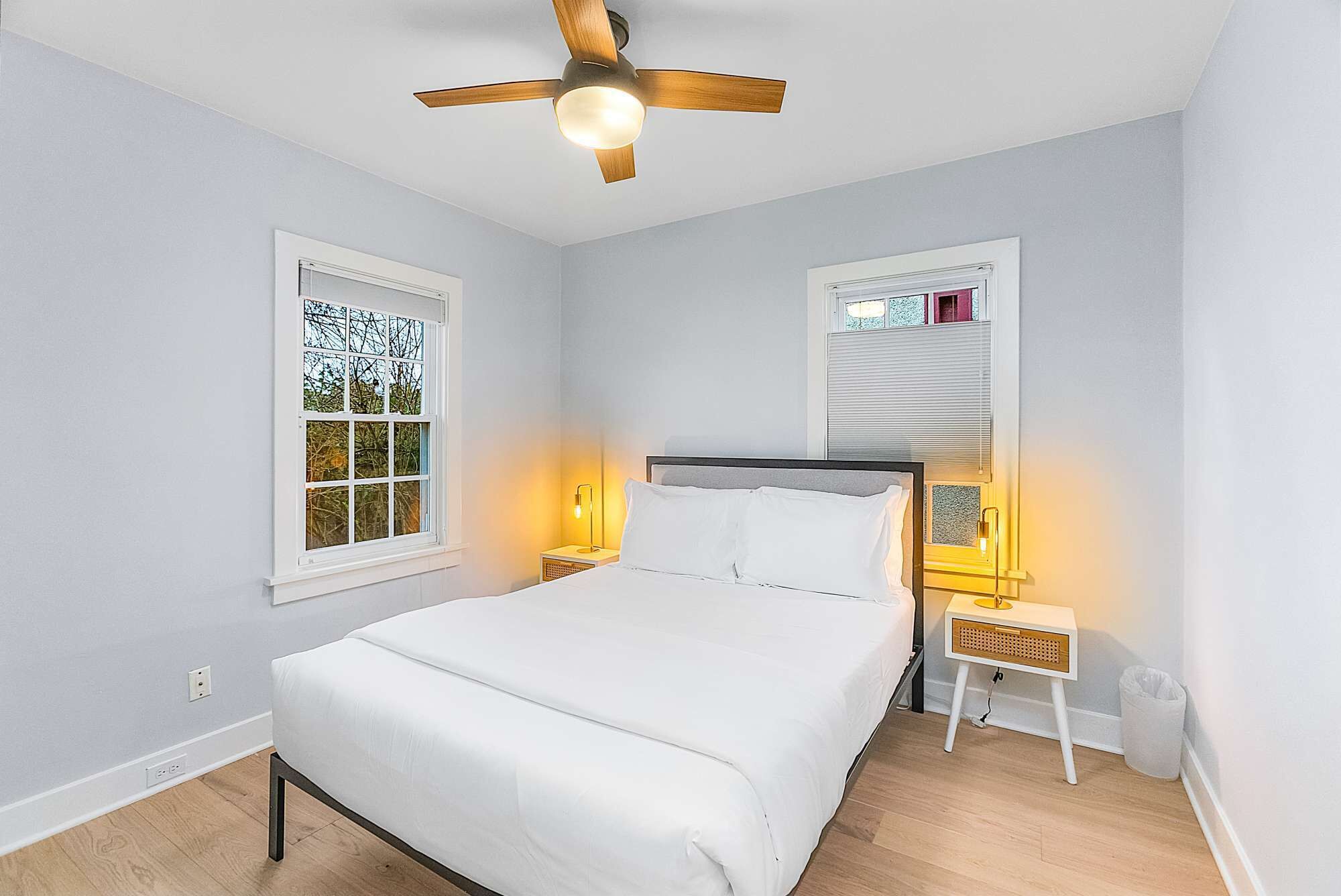 A bedroom with a bed, two bedside tables with lamps, a ceiling fan, and two windows with blinds.