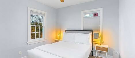 A bedroom with a bed, two bedside tables with lamps, a ceiling fan, and two windows with blinds.