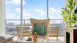 Uma varanda com vista para a cidade, uma cadeira de vime com almofada verde, uma mesa pequena com um vaso e uma estatueta, e uma planta em vaso.