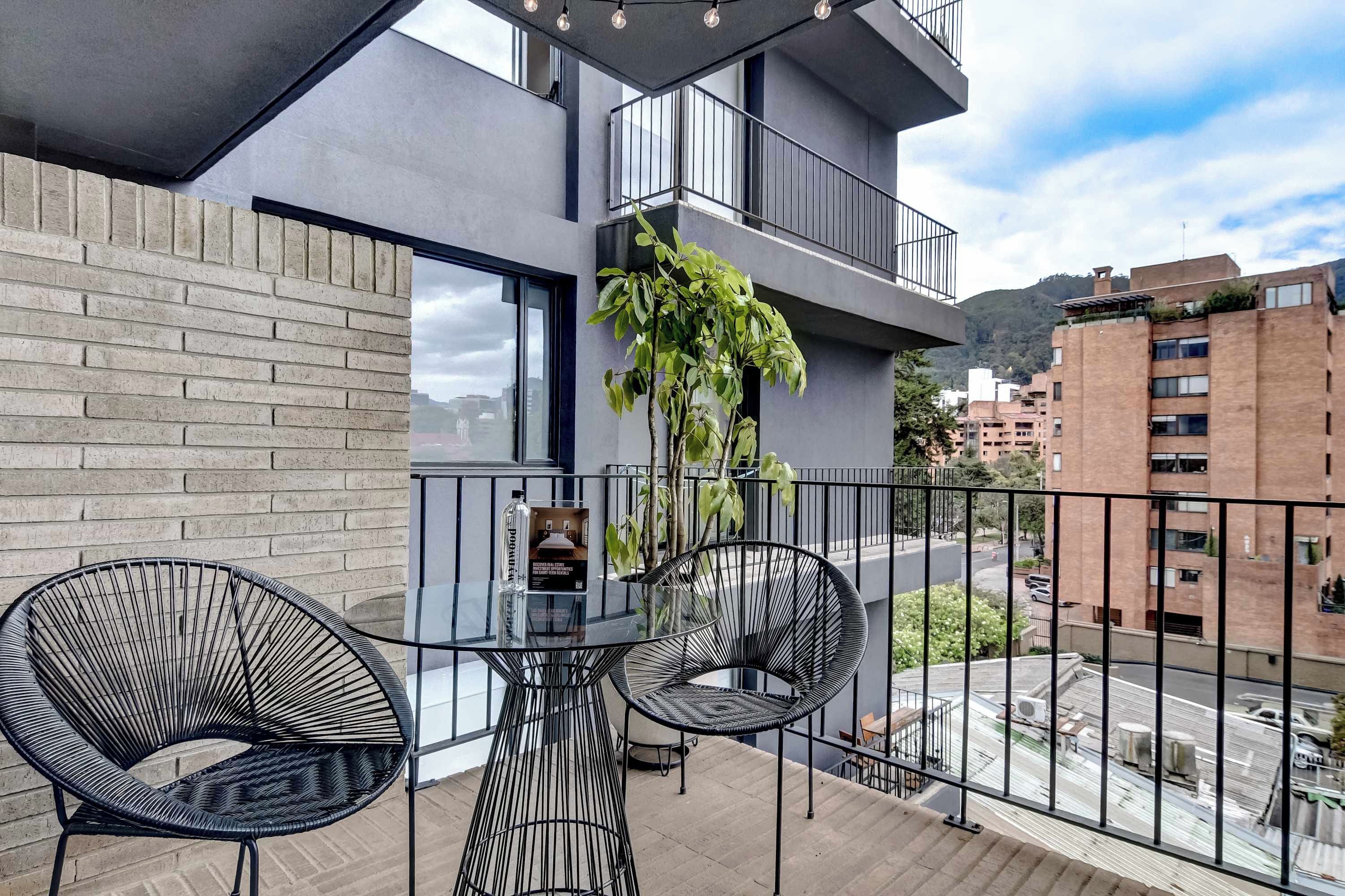 A balcony with two black wire chairs and a glass table, overlooking a street with buildings and a mountain in the background.