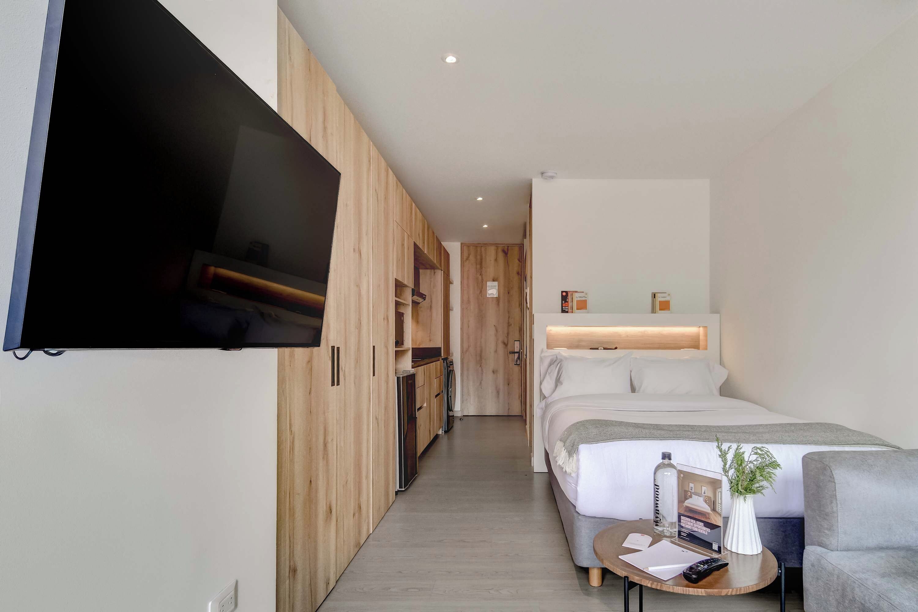 A modern hotel room with a flat-screen TV mounted on the wall, a bed with a wooden headboard, a small round table with a remote control, and a potted plant.