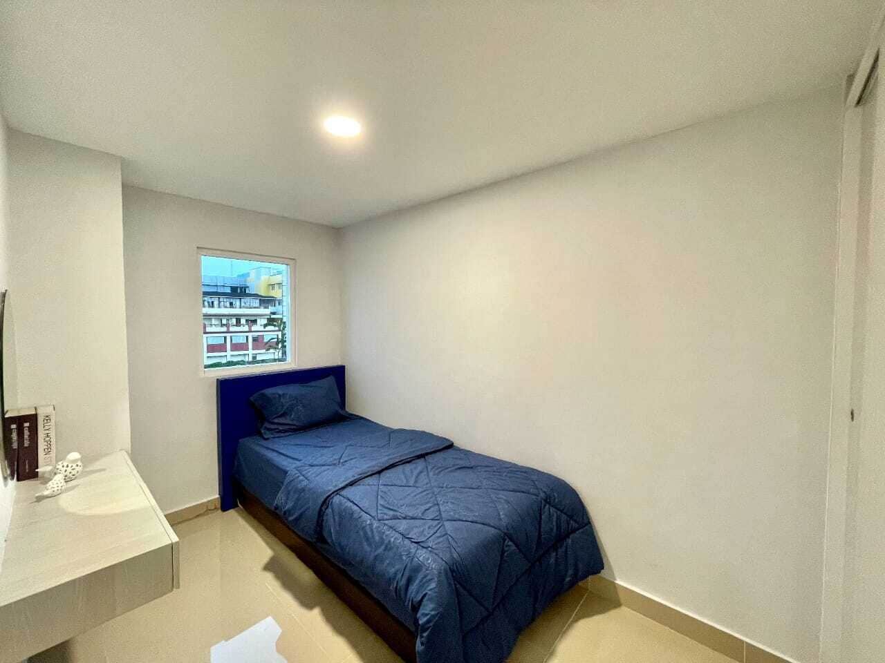 A small, single-bed room with a blue bedspread, a white sink, and a window with a view of buildings.