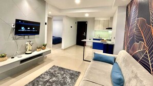 A modern living room with a flat-screen TV, a sofa with blue cushions, a grey rug, and a kitchen area in the background. -  Alhesa Apartment Mansyur Residence (Medan)