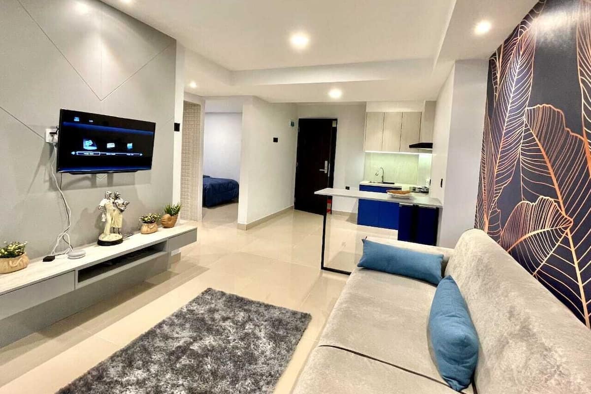 Una sala de estar moderna con un televisor de pantalla plana, un sofá con cojines azules, una alfombra gris y una cocina al fondo.