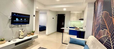Una sala de estar moderna con un televisor de pantalla plana, un sofá con cojines azules, una alfombra gris y una cocina al fondo.