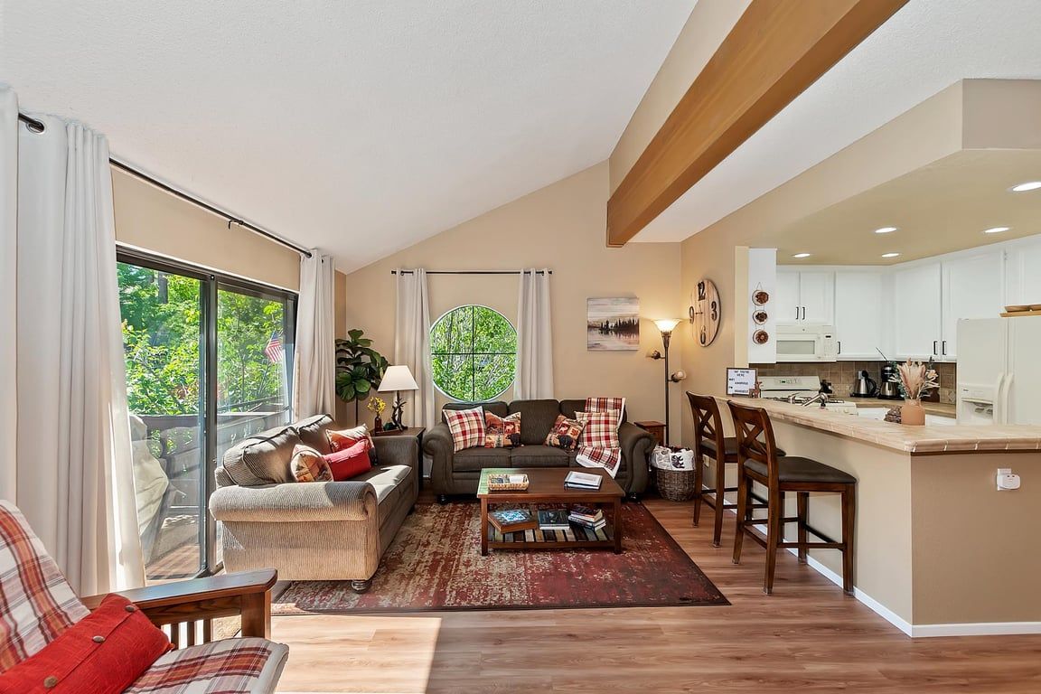 A modern living room with a sofa, coffee table, and chairs, a kitchen with white cabinets, and a large window with curtains.