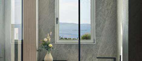 Ett modernt badrum med ett fristående badkar, ett fönster med utsikt över havet och en vas med blommor på ett litet bord i trä.