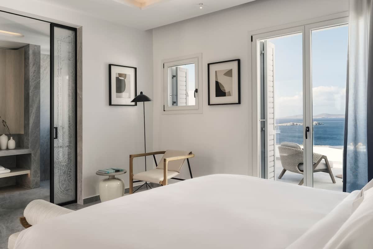 Une chambre d’hôtel moderne dotée d’un grand lit, d’un bureau, d’une chaise et offrant une vue sur la mer grâce à une porte-fenêtre coulissante.