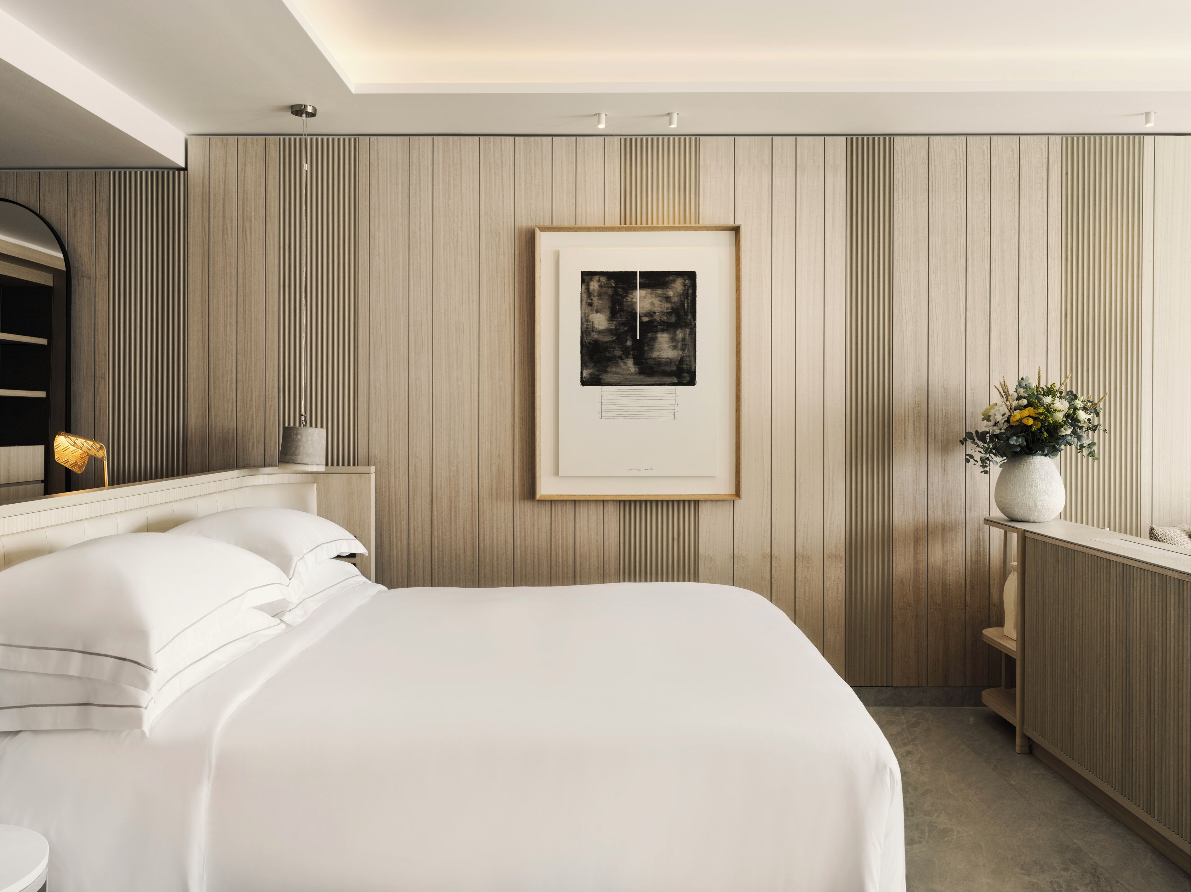 Una habitación de hotel moderna con una cama grande, una obra de arte enmarcada en la pared y un florero con flores sobre una mesita de noche.