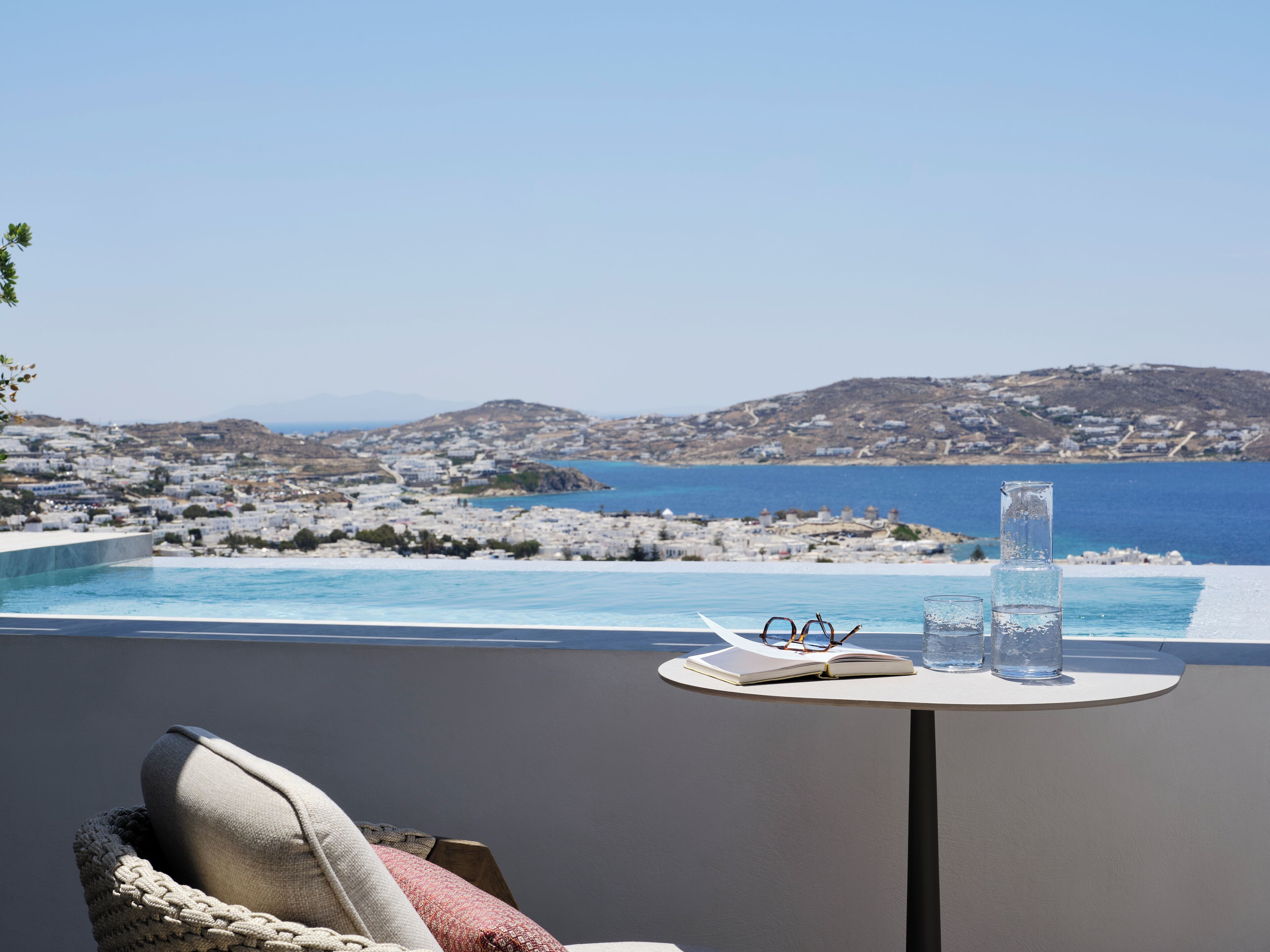 A terrace with a view of a coastal town, a pool, and a table with glasses and a book.