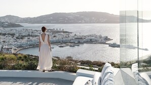 Une femme vêtue d’une robe blanche, qui surplombe une ville côtière avec ses bâtiments blancs et ses bateaux dans le port.
